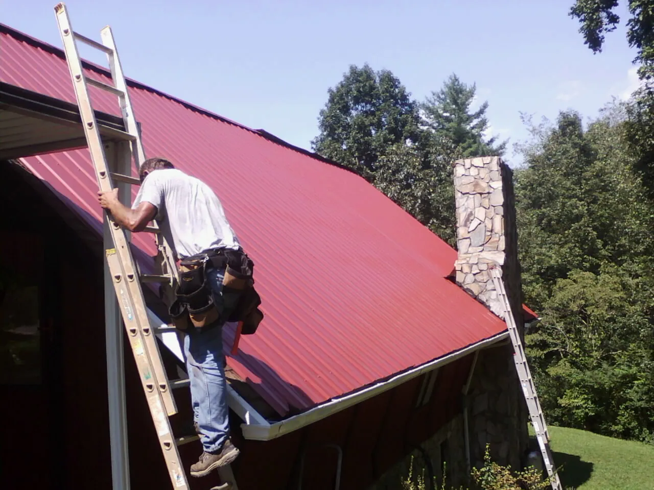 Roofer on a ladder beside a red metal roof and stone chimney — Soffit & Fascia Repair by Roofing Brothers in Little Ferry